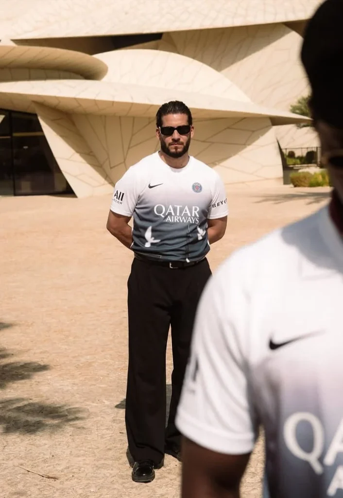 Man wearing the white-to-blue PSG pre-match jersey outside a sculptural building in Doha.