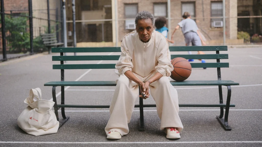 Model in cream adidas x Tyrrell Winston clothing seated on a basketball-court bench beside a ball and tote.