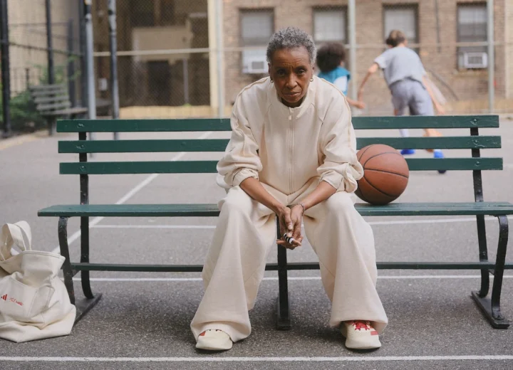 Model in cream adidas x Tyrrell Winston clothing seated on a basketball-court bench beside a ball and tote.