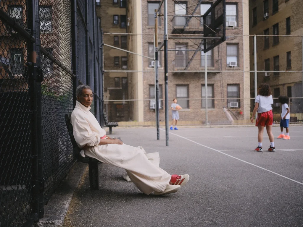 Model seated beside the fence while players use the basketball court behind.