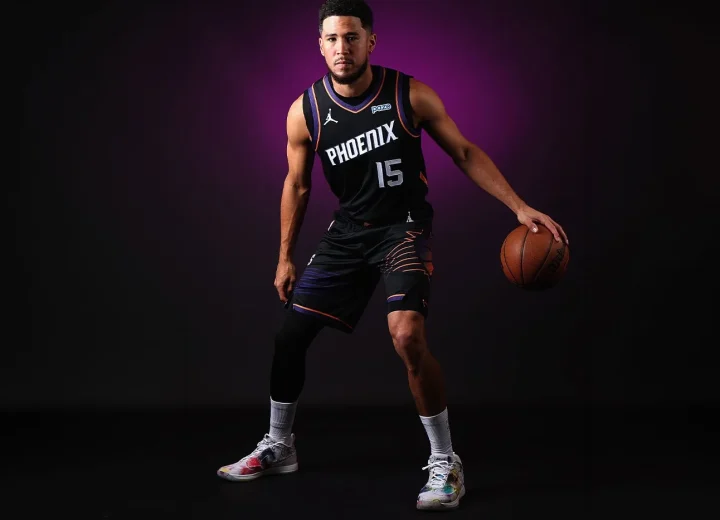 Devin Booker holding a basketball in a black Phoenix jersey while wearing Futura-designed Book 2 shoes.
