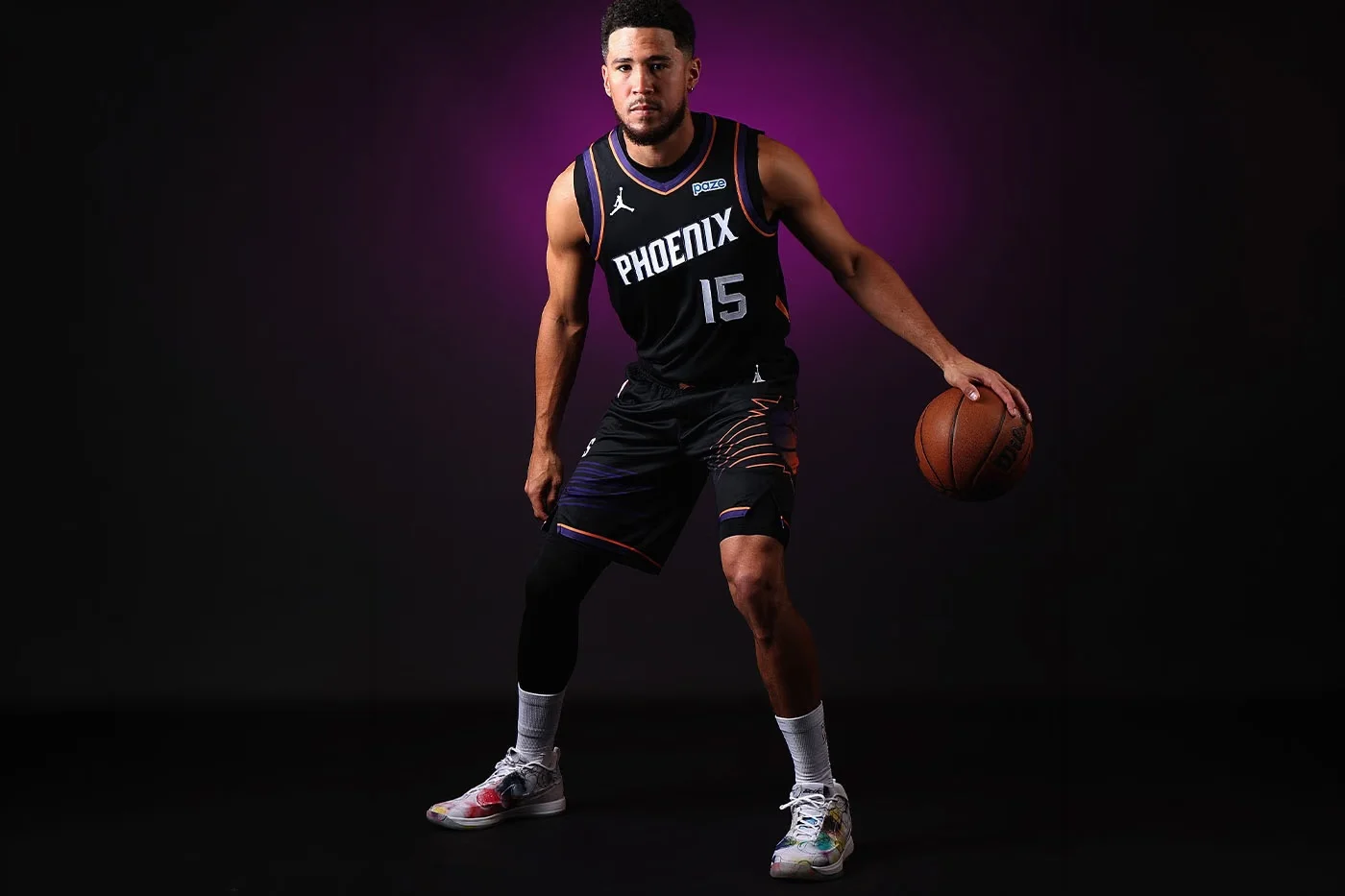 Devin Booker holding a basketball in a black Phoenix jersey while wearing Futura-designed Book 2 shoes.