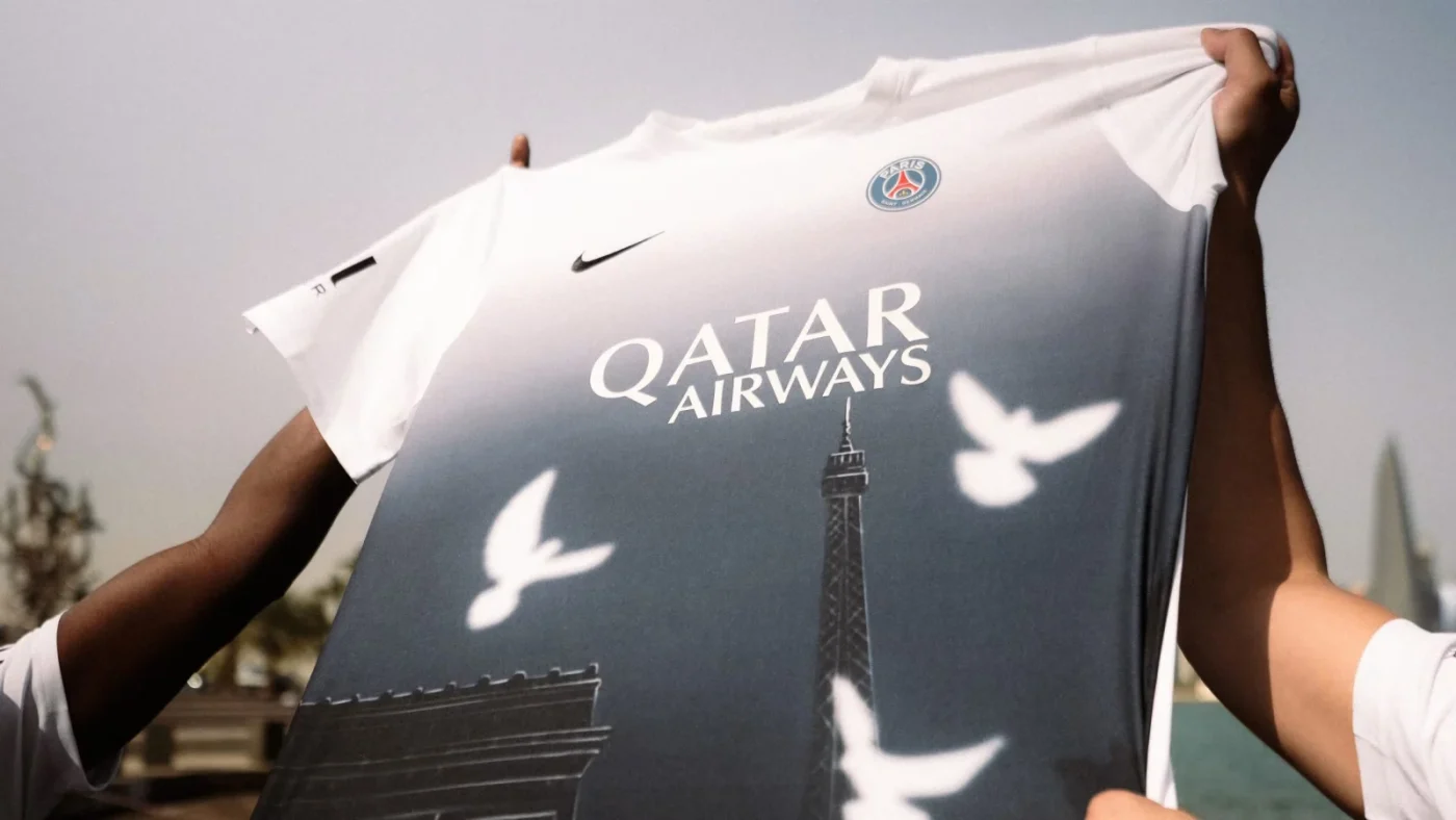 Hands hold the PSG × 3.PARADIS Art Basel Qatar pre-match shirt, with a white-to-blue gradient, white doves and Paris landmarks.