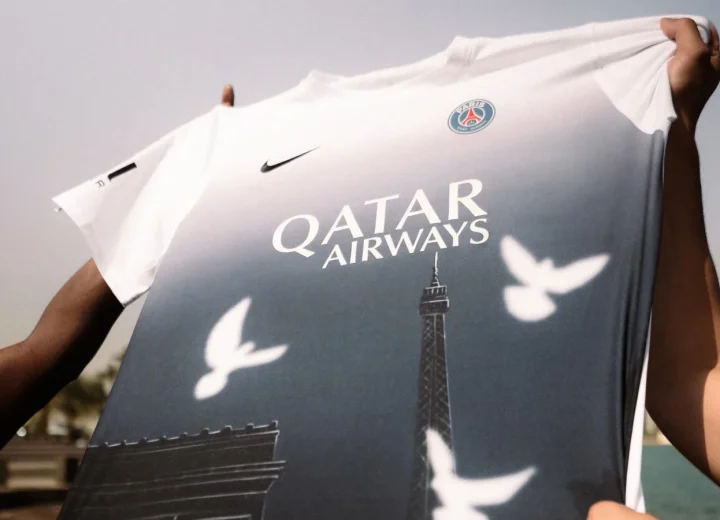 Hands hold the PSG × 3.PARADIS Art Basel Qatar pre-match shirt, with a white-to-blue gradient, white doves and Paris landmarks.
